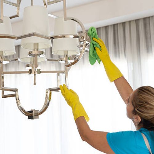 Woman worker in rubber gloves wiping dust from a contemporary ceiling chandelier