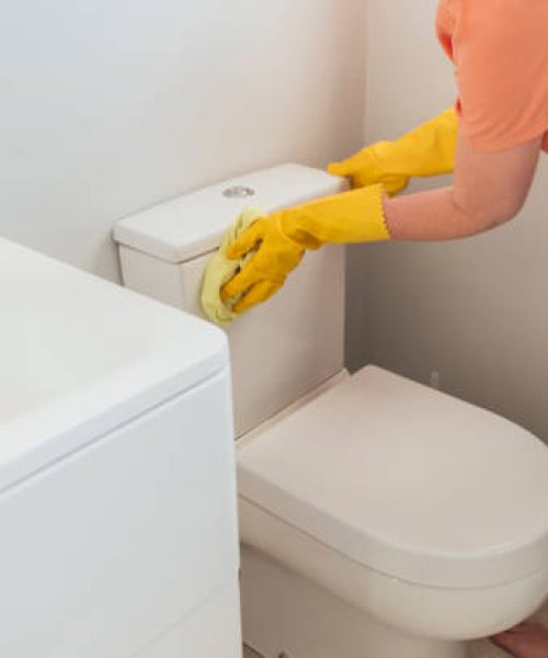 An woman worker wearing protective yellow gloves is cleaning the toilet with a sponge in bathroom.