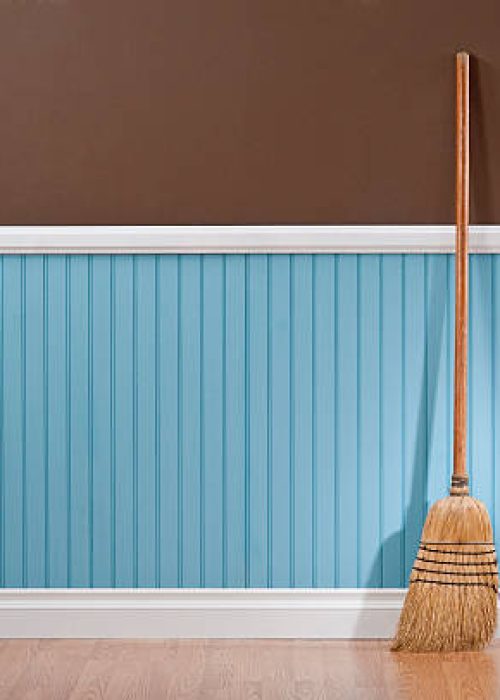 A wooden broom propped up against a wall.  The wall is two different colors.  The top half of the wall is solid chocolate brown.  The bottom half of the wall is bead board, and it has repeating parallel lines spaced a few inches apart.  The bottom section of the wall is a light shade of blue.  Propped up against the wall is a tan wooden broom.  The broom has angled bristles gathered with a dark thread.  There is white molding separating the two sections of the wall and at the floor, which is comprised of light-colored wood.