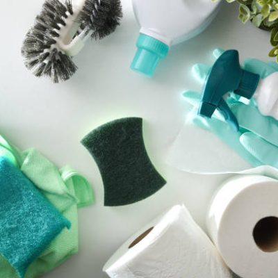 Bathroom cleaning supplies such as disinfectant bottles, scouring pads, gloves, toilet brush and toilet paper on white wooden table.