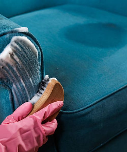 Close up of worker in pink gloves scrubbing detergent foam on upholstered armchair seat, deep cleaning fabric for home cleaning company.