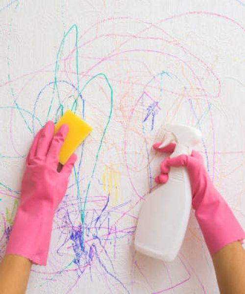 Woman hands holding detergent bottle and sponge and washing scratches lines of colorful markers from white wallpaper. Mother cleaning wall from draw of naughty little kid. Closeup. Point of view shot.